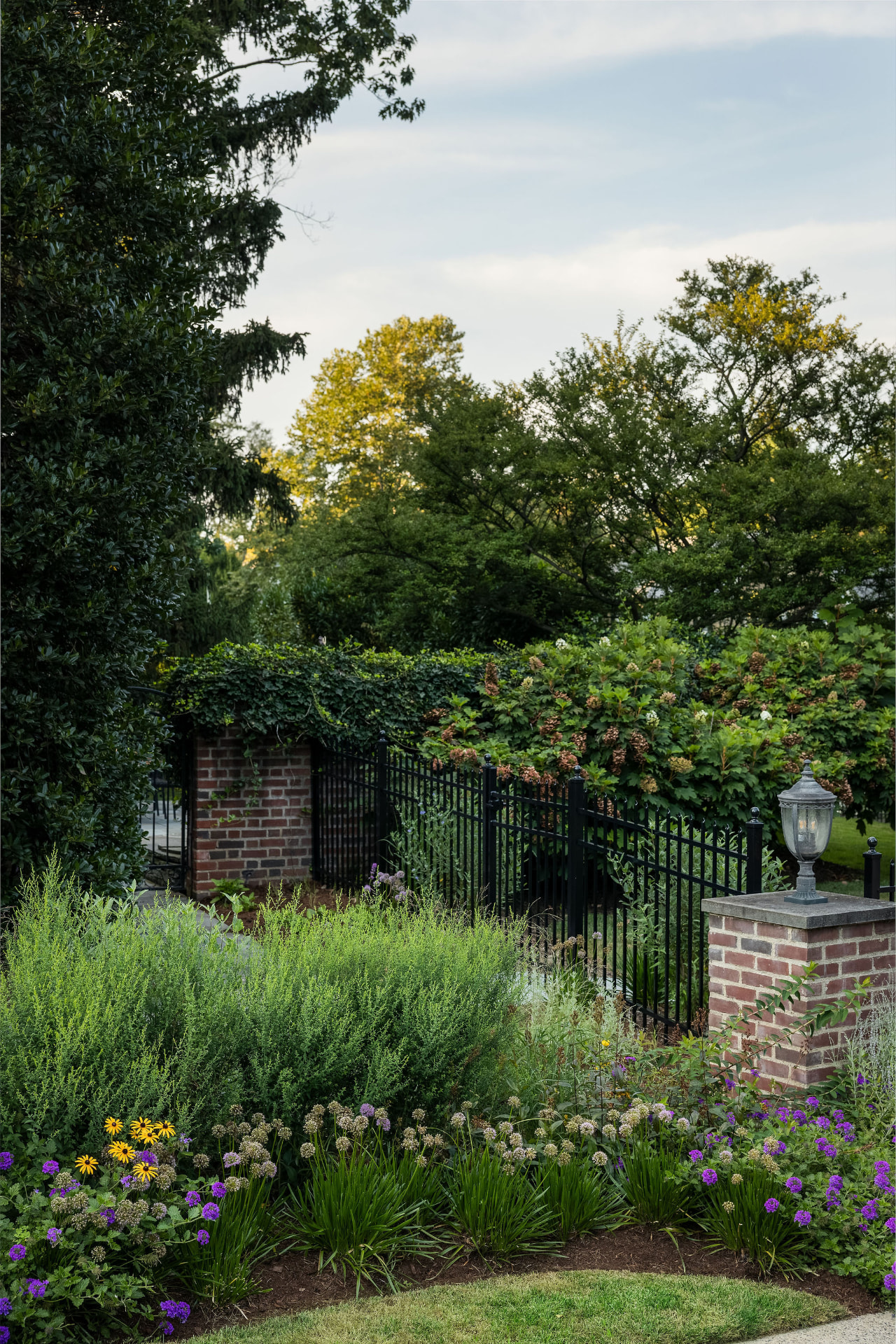 A tall view of the Derwen Road entry garden with black metal fence, brick pillars, and mature trees