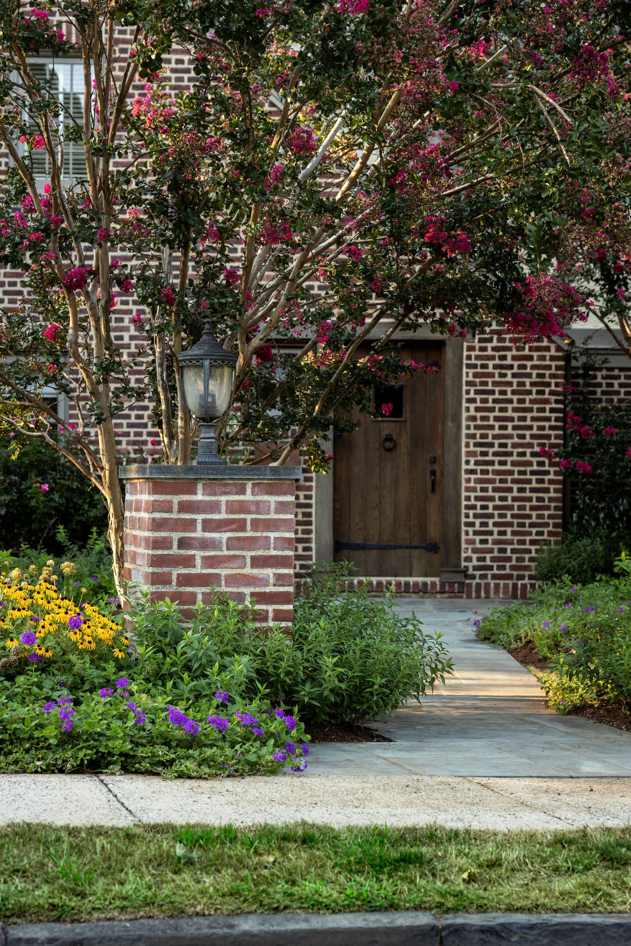 The wood front door of the brick entry building framed by crape myrtle and bluestone walk
