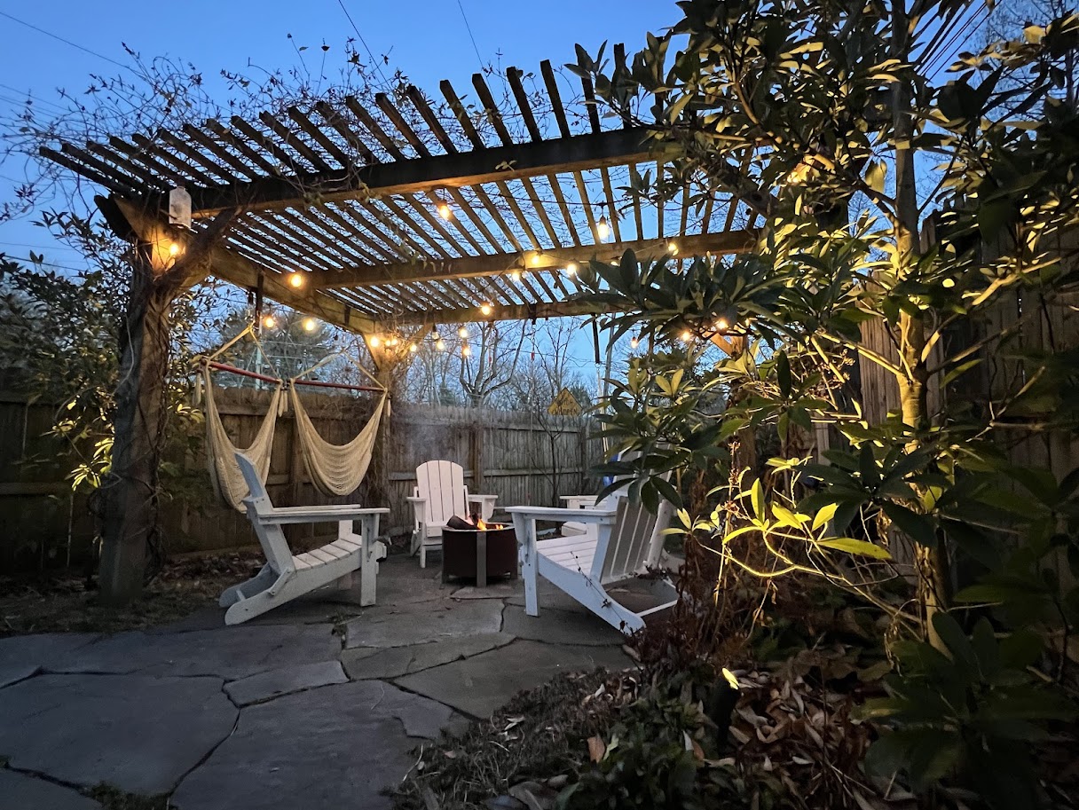 Backyard pergola with string lights and white Adirondack chairs at dusk